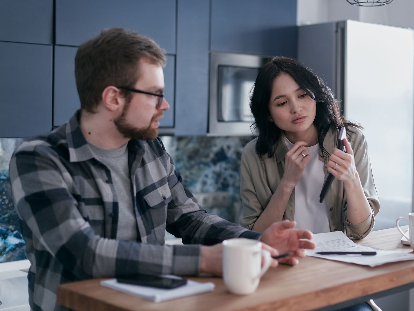 couple at opposite ends of a sofa in low kitchen light, one holding a phone / sec-1: two coffee cups on a kitchen counter in morning light / sec-2: woman's hand holding a pen over a notebook at a night kitchen table