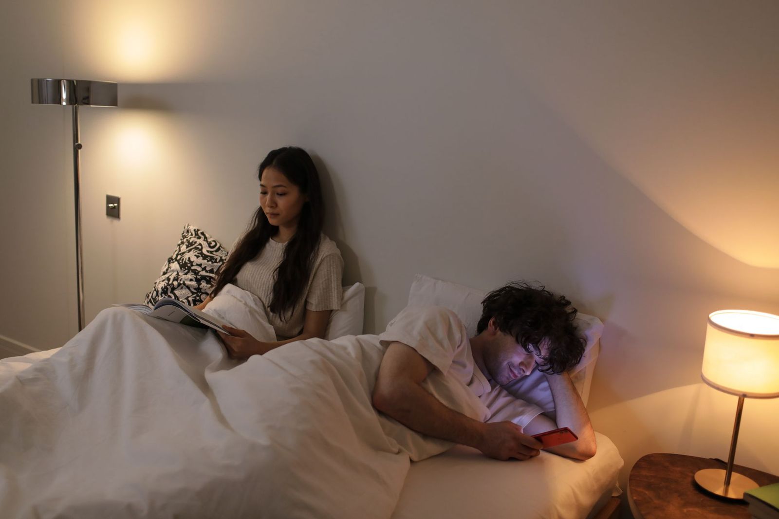 woman her thirties sitting the edge bed phone hand looking sideways man reading bed the moment before deciding alternatives woman bed holding phone beside sleeping reading man bedroom with one lit phone screen