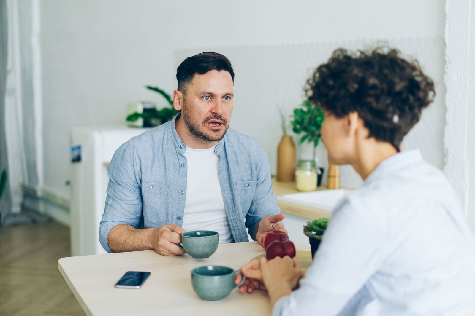 couple their late thirties kitchen table with two coffee mugs one holds phone the other handwritten list light mid conversation