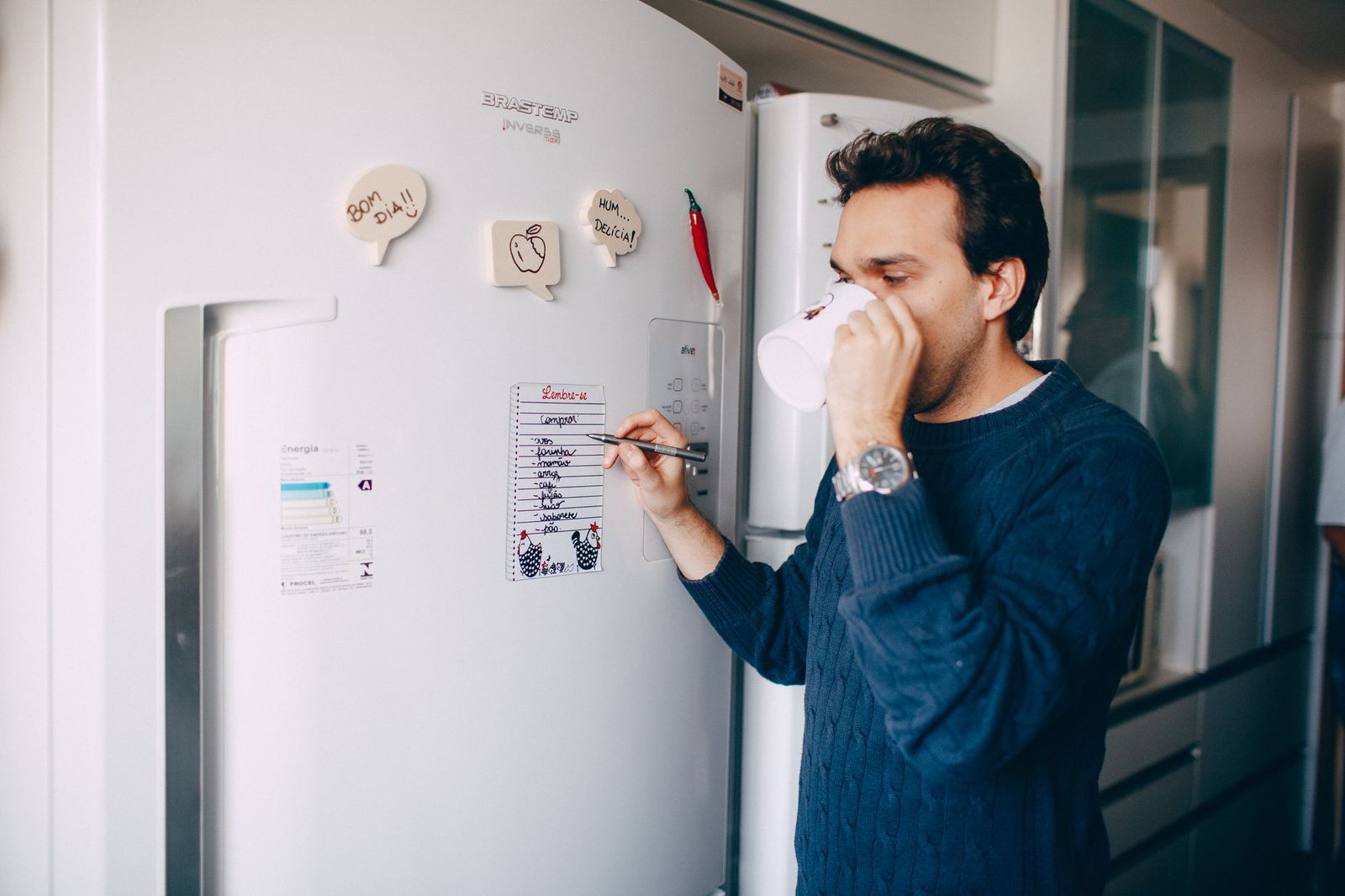 a fridge door with a handwritten list held by a magnet, a coffee cup on the counter below || fridge door with handwritten list held magnet coffee cup the counter below
