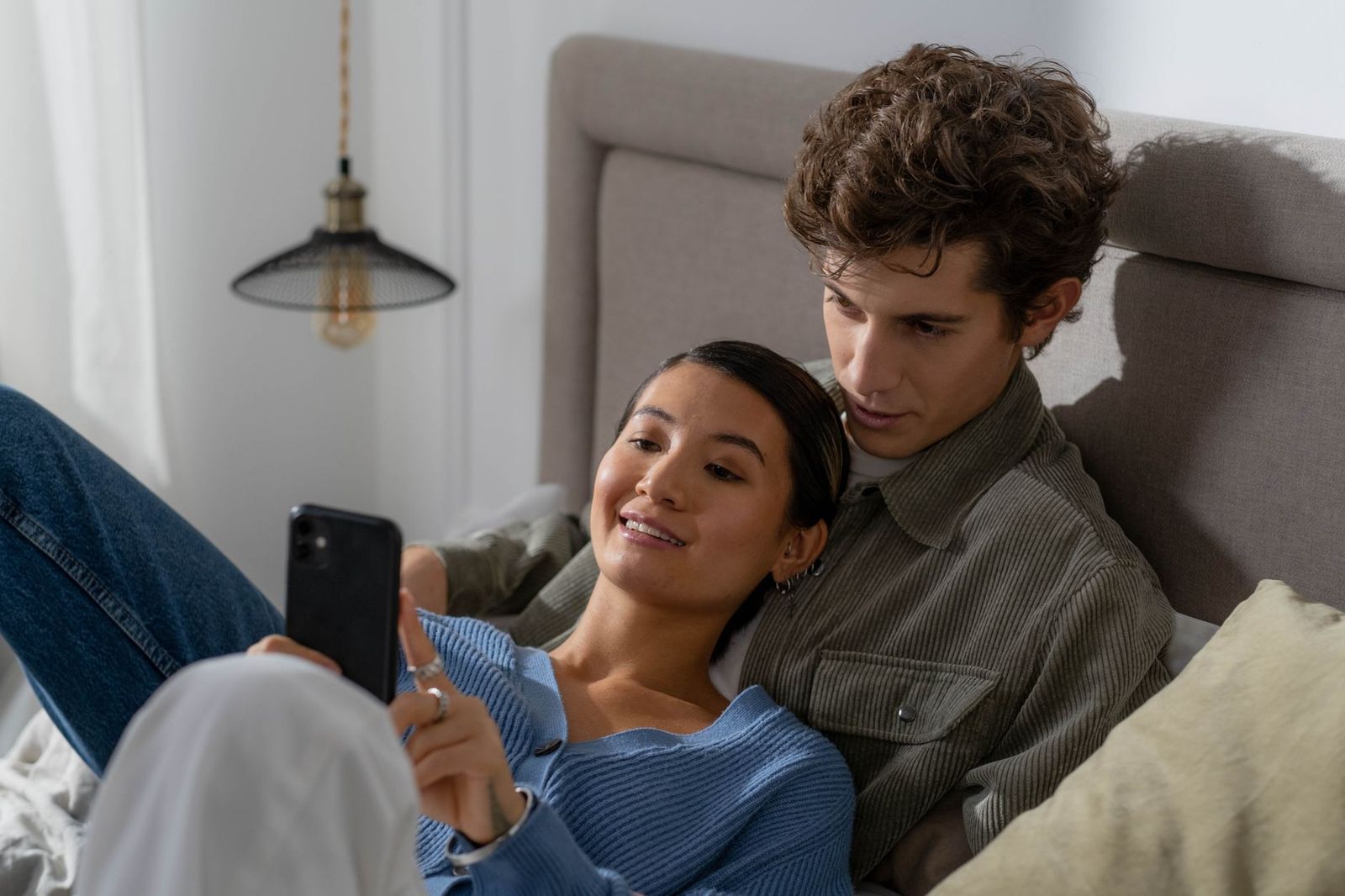 a couple sitting together on edge of bed, one holding a phone, serious warm faces || couple sitting together edge bed one holding phone faces thirties